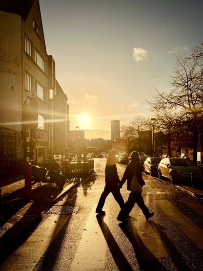 Two figures walk hand-in-hand across a zebra crosswalk at golden hour, silhouetted against a bright sun that casts long shadows on the wet street. A building stands on the left, a scooter is parked, and cars line the right side with bare trees. The vibrant sunlight creates strong contrasts and lens flare, illuminating a distant cylindrical building. The scene evokes a warm, atmospheric mood with deep shadows and bright light.