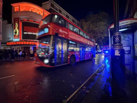 A busy city street at night featuring a red double-decker bus in motion, illuminated by nearby neon lights from a theater advertising "Stranger Things." Pedestrians walk along the pavement, and a phone booth is visible on the side. The wet road reflects the blue light from a nearby police car visible in the distance. The sky is dark