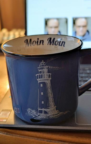 Eine blaue Emaille-Tasse mit dem Aufdruck eines weißen Leuchtturms steht auf einem Laptop. Am inneren Rand der Tasse ist der Schriftzug "Moin Moin" zu lesen.