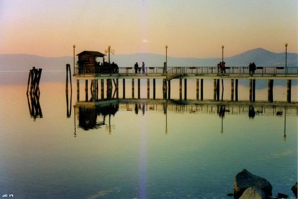 Farbfoto, aufgenommen bei Sonnenuntergang am Lago di Bracciano in Italien. Ein Fußgängersteg führt von rechts nach links in den See und wird im Wasser reflektiert. Am Horizont dunstig die umliegenden Berge. Darüber der Himmel in weichen Orangetönen. Leica R3 auf Kodak-Film.