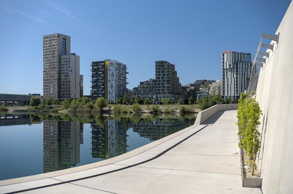 Das Foto zeigt eine moderne, urbane Szene an einem sonnigen Tag. Im Vordergrund sieht man eine Promenade, die entlang eines ruhigen Sees verläuft. Das Wasser spiegelt die umliegenden Gebäude wider, was einen ästhetischen Effekt erzeugt.
Im Hintergrund sind mehrere moderne Hochhäuser und Wohngebäude zu sehen, die in verschiedenen architektonischen Stilen gestaltet sind. Einige Gebäude haben eine glatte, klare Fassade, während andere durch ihre ungewöhnlichen Formen auffallen. Die Umgebung wirkt sauber und gepflegt, mit etwas Grünfläche und Bäumen, die zwischen den Gebäuden verteilt sind. Der Himmel ist klar und blau, was die moderne Atmosphäre der Szene unterstreicht.