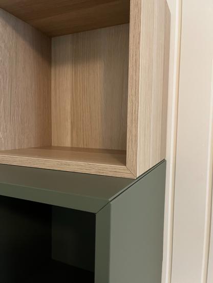 Houten en donkergroene kast. Het bovenste deel is gemaakt van licht hout met een open vak, en daaronder zit een donkergroen metalen of houten gedeelte, ook met een open vak. De stijl is modern en strak, met scherpe rechte hoeken. De kast staat tegen een lichte muur.