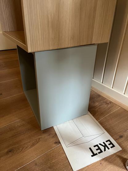 Een kastje of meubel met een houten bovenkant, geplaatst op een houten vloer. Onder het kastje ligt een papieren handleiding waarop een 3D-tekening van het meubel staat afgebeeld. Op de handleiding staat groot het woord "EKET", wat waarschijnlijk de naam van het product of een modelnaam is. De handleiding is deels onder het meubel geklemd.