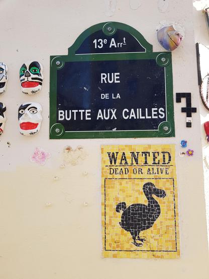 A black on yellow mosaic of a Wanted Dead or Alive poster featuring a dodo. It's positioned bellow a "Rue de la Butte aux Cailles" street name plaque next to a bunch of other small art pieces.