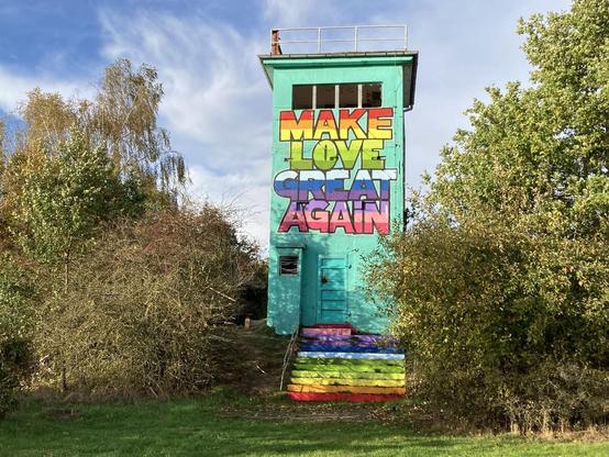 Helles, grünes, quadratisches Gebäude mit bunt gestreifter Fassade und Aufschrift "MAKE LOVE GREAT AGAIN" im Freien, umgeben von Gras und Büschen.