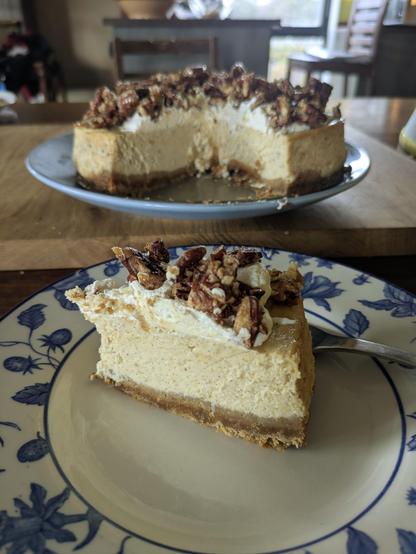pale cream cheesecake slice on a blue and white dessert plate. the rest of the cheesecake in the background. 
the cake has a dark brown biscuit base, a thick creamy coloured cheese layer and dark brown brittle on cream topping.
