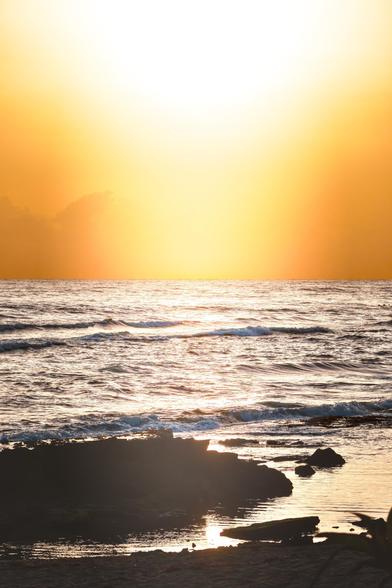 A serene coastal scene featuring the sun setting on the horizon, casting an orange glow over the water. Gentle waves lap at the shore, with dark rock silhouettes in the foreground.