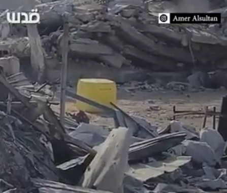 Cropped screenshot of a rubble strewn landscape of buildings razed to the ground.

In a path between rubble in the foreground and rubble in the background stands a yellow concrete cube. One of Israel's markers indicating that they'll shoot to kill any living thing (human or animal) who moves past it.

Station and program identification logos appear in the upper right- and left-hand corners, reflecting the fact that this has been taken from some form of media agency.