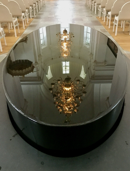 Das steinerne, ovale Taufbecken im Mittelgang der Kirche hat eine spiegelglatte Wasseroberfläche. Darin spiegelt sich das helle, barocke Deckengewölbe, der Deckel der Kanzel und ein vielarmiger Kronleuchter aus Messing. Zu sehen ist auch ein heller Holzfußboden und weiße Stühle in Reihen. (Ja, diese Kirche gefällt mir sehr!)