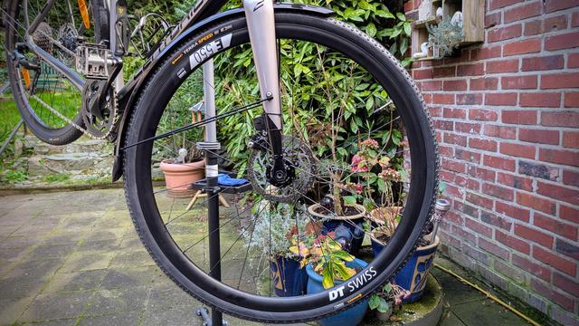 Ein silber-graues CENTURION-Allroadbike auf einem Montageständer. Die Bildmitte wird vom (neuen) Vorderrad ausgefüllt, darauf zu lesen sind "DT SWISS" und "E550", auf dem Reifen "Continental" und "Terra Speed".
Rechts ist eine rote Ziegelmauer, im Hintergrund das Grün eines Gartens.