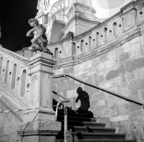 The black-and-white photograph captures a stone staircase on the grounds of St. George’s Cathedral in Lviv. The scene is illuminated at night, revealing ornate architectural details — curved balustrades, sculpted figures, and textured stone walls. A weathered statue of a cherub stands atop the railing in the foreground, while its shadow is dramatically cast onto the steps below, creating a sense of quiet mystery and depth. The play of light and shadow emphasizes the baroque elegance of the setting and evokes a contemplative, almost cinematic atmosphere.