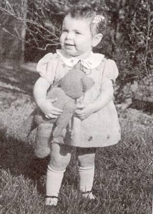 A very young girl in a dress standing in a garden. She is holding a large teddy bear in her hands.