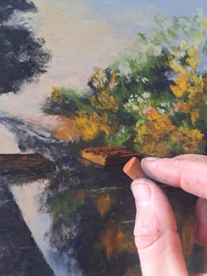 Painting of a canal with trees in autumnal colors. Fingers holding a brownish soft pastel as if painting with it.