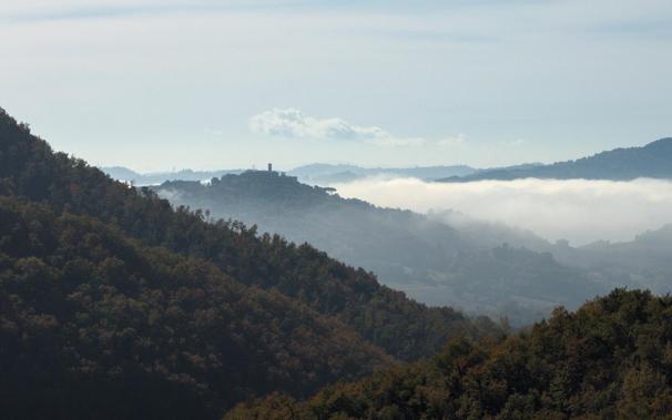 A view of rolling hills covered by Mediterranean woods. In the centre a small village with a prominent tower sits on a hill that rises above the low clouds and haze in the valleys.