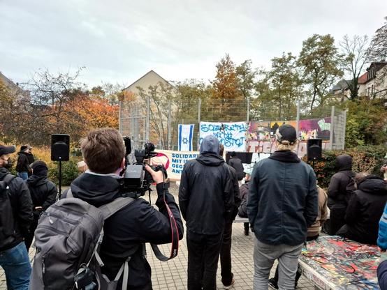 Demonstration: Die meisten Personen tragen dunkle Kleidung, darunter eine Person mit Kapuzenpullover und eine andere mit einer dunklen Kappe im Baseballstil. Links und rechts im mittleren Bildbereich sind kleine schwarze Lautsprecher aufgestellt. Am Zaun im Hintergrund sind Graffiti und zwei Plakate zu sehen. Das Plakat auf der linken Seite zeigt eine hellblaue Fläche mit einem weißen Davidstern, das an eine stilisierte israelische Flagge erinnert. Rechts daneben ist ein Plakat, das mit großen, blauen und geschwungenen Buchstaben die Aufschrift „SOLIDARITÄT MIT ISRAEL. GEGEN JEDEN ANTISEMITISMUS“ trägt. Die Mauer dahinter ist mit bunten Graffiti bedeckt. Hinter der Mauer bilden Herbstbäume und ein graues Haus den Hintergrund, darüber ein heller, bewölkter Himmel. Die Beleuchtung ist weich, was auf einen bewölkten Tag hindeutet.
​Englische Zusammenfassung: An outdoor, eye-level shot of a group of people facing a wall with posters and graffiti, including one with a blue and white Star of David design and another reading "SOLIDARITY WITH ISRAEL. AGAINST ALL ANTISEMITISMUS". A person in the foreground is holding a video camera, suggesting a media presence at an event or demonstration. The scene is set in an urban area with autumn foliage.