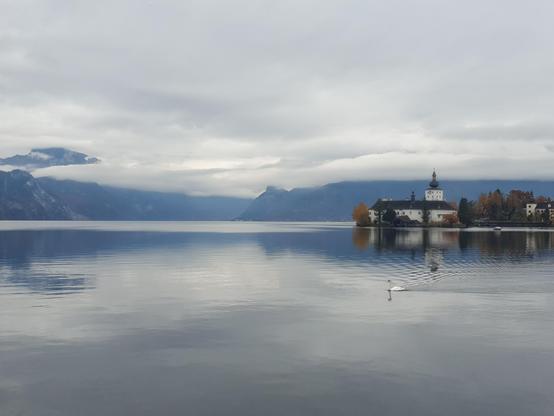 Spuegelglatter See, in dem sich Berge und tiefhängende Wolken spiegeln. Rechts im Bild ist ein kleines Wasserschloss.