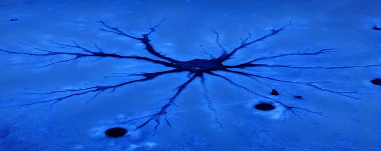 At twilight a melted patch on the thinning lake ice is a deep blue against the lighter blue of the ice, with long, branching tentacles of melting extending out in every direction.