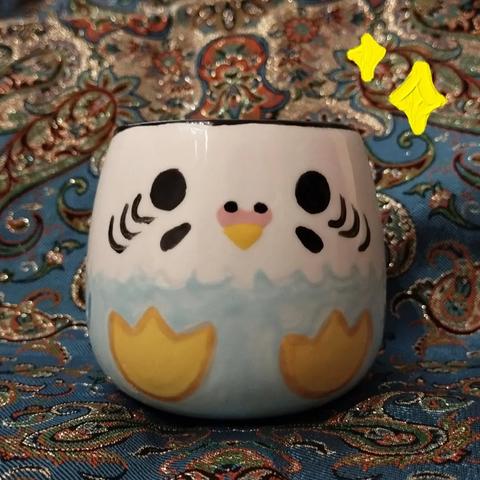 Photo d'une tasse en céramique. J'ai peins dessus une perruche ondulée bleue, sur toute la tasse, comme si la tasse était la perruche. Elle est mignonne. Elle est posée sur un termeh, une nappe iranienne aux riches décors bleu et or.