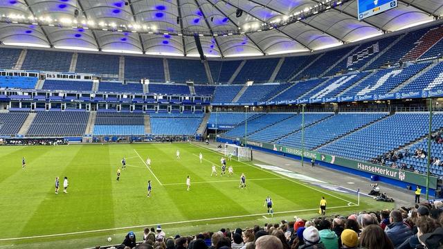 Blick auf eine Hälfte in einem Fußballstadion, wo der Ball gerade auf dem Weg Richtung 16-Meter-Raum ist, die Ränge im Hintergrund sind leer.