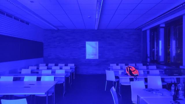 Ein Foto eines sehr tief-blau ausgeleuchteten Klassenzimmers. Auf einem der Tische liegt ein strahlend neon-orangener Rucksack.