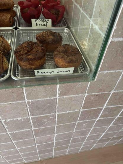 Un coin de vitrine dans une pâtisserie, en haut à gauche. Le reste de la photo montre un mur carrelé de carreaux rose pâle.
Dans la vitrine, au premier plan, 3 kouign amann avec une étiquette indiquant ce mot. Derrière, d’autres gâteaux qui ressemblent à des muffins avec une étiquette « Monkey bread »