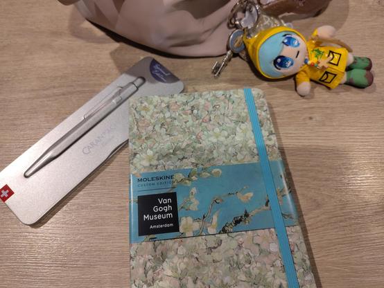 Veganes Moleskine-Notizbuch als van-Gogh-Sonderedition, dazu ein wiederauffüllbarer Kuli made in Switzerland (Caran d'Ache, in einer Sonderfarbe). Im Hintergrund die Stoffhandtasche mit Luce, dem Vatikan-Maskottchen.