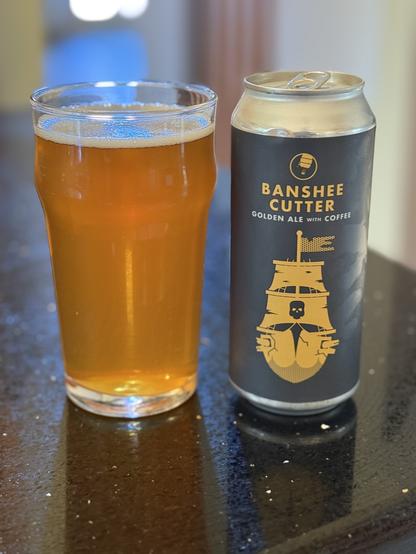 A pint glass of golden ale sits on a shiny black counter top. The black pint can has a golden pirate ship with a black skull on its taut mainsail