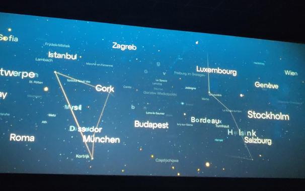 El anuncio de Europa Cinemas. Numerosos nombres de ciudades europeas aparecen en letras blancas de diferentes tamaños, destacando algunas como Sofía, Estambul, Cork, Düsseldorf, Múnich, Budapest, Luxemburgo, Ginebra, Estocolmo, Helsinki, Roma, Viena, Zagreb, Burdeos y Salzburgo. Finas líneas blancas conectan algunas de estas ciudades, formando figuras geométricas que recuerdan a constelaciones, como un triángulo prominente que une Cork, Düsseldorf y Múnich, y una línea que conecta Luxemburgo, Burdeos, Helsinki y Salzburgo.