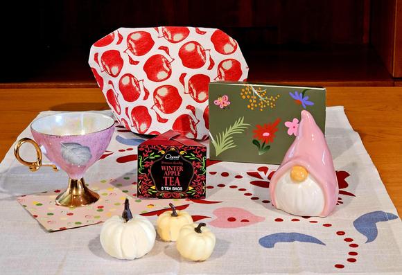 Tea tableau on snowy white linen decorated with a soft pink, red, and blue Scandinavian design. There is a white tea cozy with dusky red apples, a muted green art card with flowers and ferns, a pink ceramic gnome, three miniature white pumpkins, a tall, delicate pink cup with ornate gold handle and pedestal, with a fad3d green leaf handpainted on it, and a small triangular red and green box of Winter Apple black tea, both of which sit upon a pink napkin with a pattern of tiny green, red, and ivory apples.
