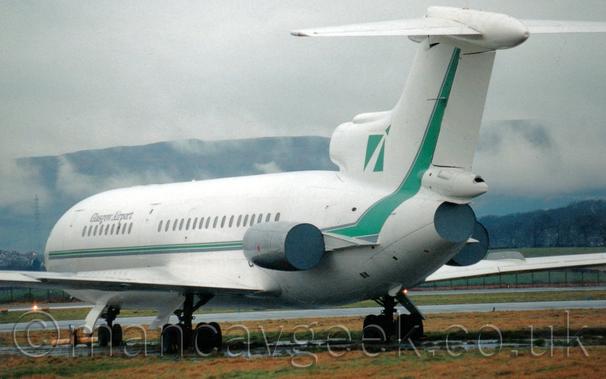 Rear view of a 3 engined jet airliner, with the engines removed but the engine pods left in place, parked facing to the left and slightly away from the camera on concrete pads in the middle of a grass field.
The plane is almost entirely white, with a green stripe running along the body, sweeping up into the tail, and black "Glasgow Airport" titles on the upper forward fuselage.
Grey metal plates cover the engine exhausts.
Hills rise up in the distance, partially obscured by smears of grey, low lying cloud, with flat grey cloud beyond that.