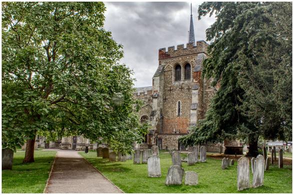A historic stone church with a square tower and small spire stands among tall trees and old gravestones under a grey, clouded sky. A narrow path leads toward the church entrance, framed by lush green foliage and weathered headstones, giving the scene a peaceful yet timeworn atmosphere.