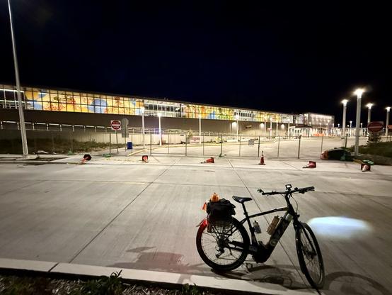 My e-bike sits on the side of a road with its lights on in front of the future Star Lake light rail station which has colorful murals on the elevated train platform