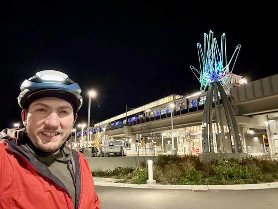 Selfie smiling in front of a lit up metal sculpture of abstract hands in front of the elevated Federal Way Downtown light rail station as a light rail train undergoes a test run at the train platform