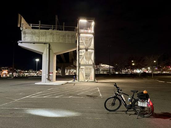 My e-bike with its lights on sits on the ground in front of the southern end of the elevated light rail tracks where a concrete support ends