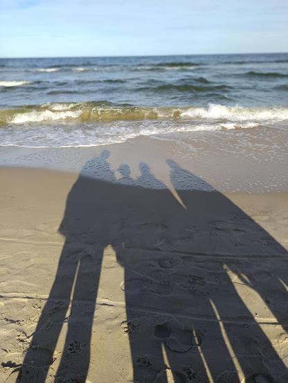 Four shadows of people. They are two long shadows left and right, the two in the middle are shorter. The shadows are on the beach, at the end of the pic you see the sea and the sky. It's a sunny day.
