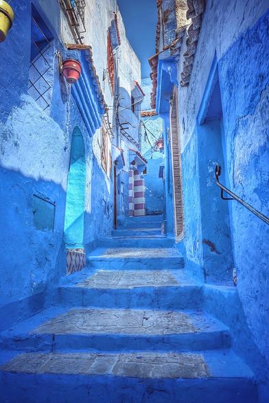 A highly saturated, low-angle photograph of a narrow, steep alleyway in Chefchaouen, Morocco. The walls and steps are painted a striking, deep blue. Sunlight casts strong shadows, highlighting the textured stone and uneven steps leading up into the light. A hint of an arched doorway and a distinct red lantern are visible on the left side of the narrow passage.