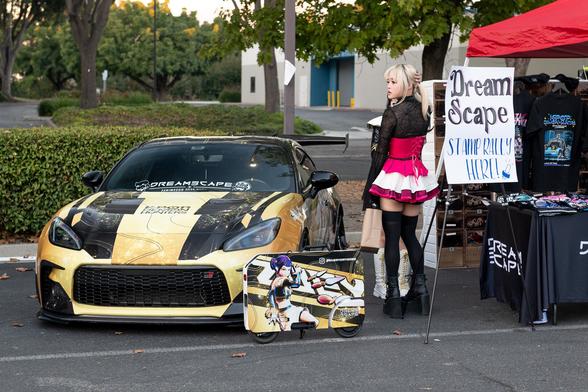 A blonde-haired girl wearing a black top, fuchsia corseted skirt, and black thigh-high stockings and tall boots, standing next to a gold-and-black wrapped GR86 sports coupe, and a K-Pop Demon Hunters-skinned Motocompacto scooter. A booth next to them has a sign that reads “Dream Scape Stamp Rally Here!”