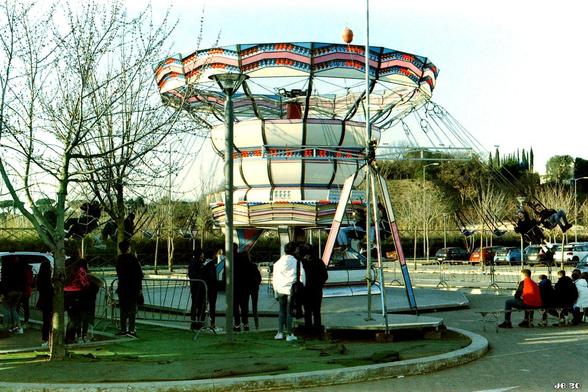 Farbfoto eines sich in voller Fahrt drehenden Kettenkarussells auf einem Platz in Rom. Im Vordergrund stehen und sitzen etliche junge Zuschauer. Etwas trübe Farben wegen des verwendeten Films (Lomochrome Metropolis).