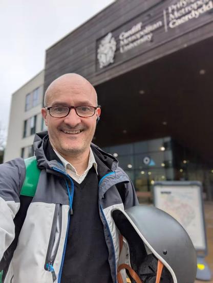 A bald man wears glasses and holds a bicycle helmet in his hand. He is standing in front of a blurred building which has the name 'Cardiff Met University' on it.