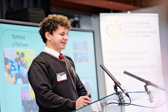 A young man in school uniform stands at a lectern in front of a screen and a pop-up stand.