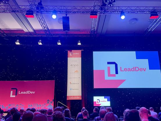 A conference stage with the LeadDev logo shown on 2 big displays. A crowd sitting in front of the stage. Colours are in the pink and blue spectrums.