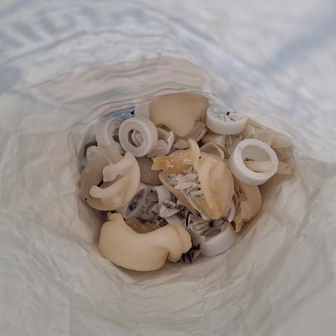 Blick in eine zerknitterte helle Papiertüte in der Reste von beigen Kerzenstumpen und weißen Teelichtern liegen. Zwischen zerbrochenen Wachsresten in unterschiedlichen Größen und Formen liegen kurz schwarze verbrannte Kerzen Dochte. Die vorherrschenden Farbtöne sind Sandgelb, Elfenbein und Weiß.