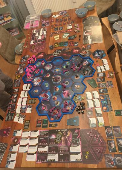 Photographs that shows a tabletop game set up on a wooden table. The central focus is a hexagonal board with a blue border, featuring various hexagonal tiles with illustrations of planets, space stations, and other sci-fi elements. The board is populated with small, colorful game pieces, including red and blue tokens, and a few larger figures.

Surrounding the board are numerous cards and tokens, some with text and symbols, indicating different game mechanics. The cards are organized in piles and stacks, with some laid out in rows. There are also dice and other game components scattered around the board.