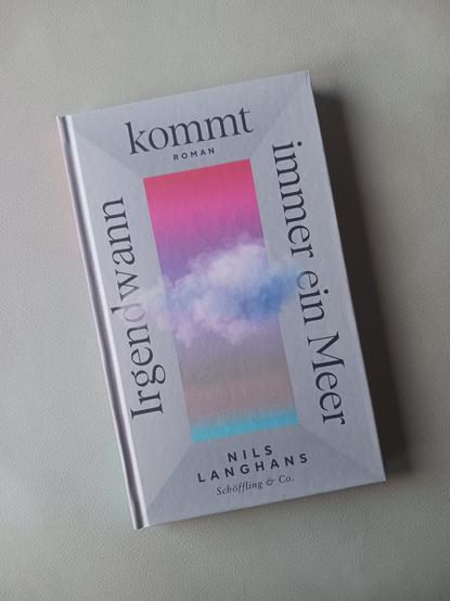 Foto des Buches, auf dessen Cover der Titel in einem grauen Rahmen um ein pink- und türkisfarbenes Rechteck läuft, aus dem eine Wolkengebilde auf den Rahmen ragt. Das Buch liegt auf grauem Hintergrund.