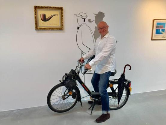 Merveille with white shirt and blue jeans posing on a velosolex in an expo room. On the wall behind him: a painting by Magritte Ceci n'est pas une pipe (not the original , which is larger) and Merveille's own drawing of Monsieur Hulot. Also his shadow was painted, where Hulot has the pipe in his mouth. Think this one through... :)