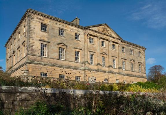 A sunlit golden sandstone stately home viewed from low down and quite close up, from behind a terrace garden wall which is at the bottom of the image. There is a blue sky.