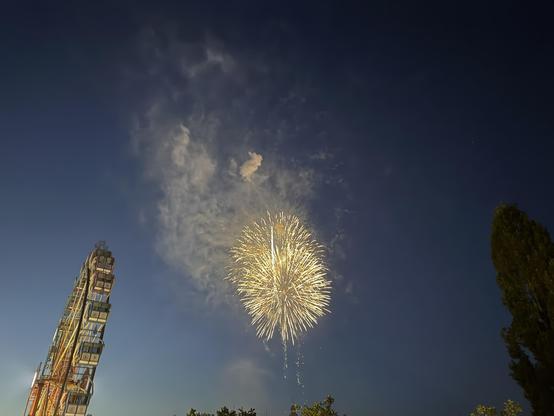 Ein lebhaftes Feuerwerk erhellt den Nachthimmel, umgeben von Rauchschwaden. Im Vordergrund ist teilweise ein Riesenrad zu sehen, das sich vor dem Hintergrund des bunten Feuerwerks abzeichnet.