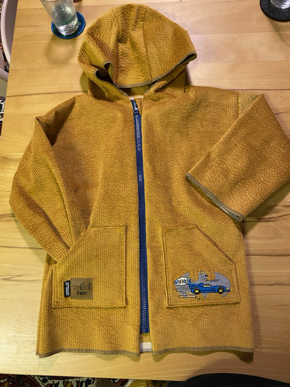 Gelbe Außenjacke aus Baumwollcord mit eckigen, aufgenähten Taschen. Auf einer Tasche ist eine Stickerei von einem Batmobil. Auf der anderen Tasche ist ein Aufnäher mit dem Wort „Attacke“ und dem Schriftzug „wild one“. Der Reißverschluss ist blau.