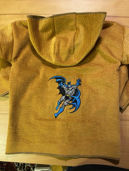 Es ist die Rückseite einer gelben Cordjacke zu sehen. Sie hat eine Kapuze und eine Stickerei auf der Batman sich an einem Seil durch die Luft schwingt.