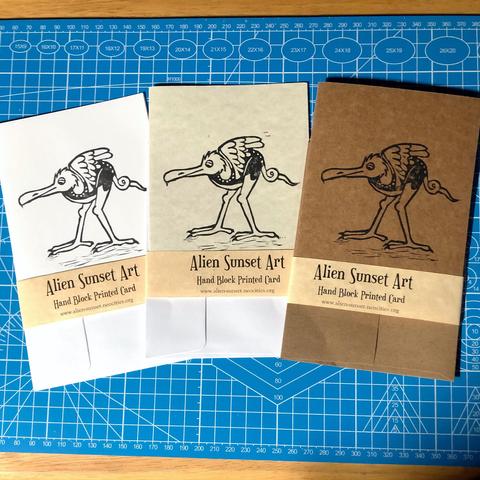 a set of three cards laid out on a blue cutting mat. a brown Kraft card, a plain white card, and a cream parchment pattered card. each card has a yellow parchment strip of paper wrapped around it that says "Alien Sunset Art" Hand Block Printed Card.

the block print on the cards is of  a long legged bird like creature with a curling tail and very long beak